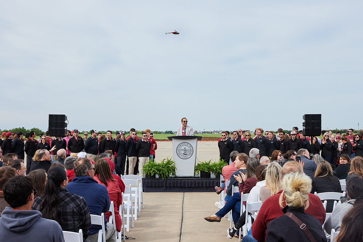 President Harroz speaks at a podium before the assembled crowd at the Aviation Fly-In event. A helicopter flies overhead.