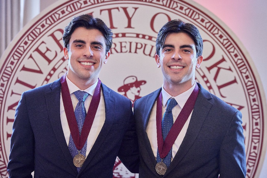 Twin brothers David and Eric Barroso posing for a photograph adorned with their 4.0 commemorative medallions in front of an OU seal.