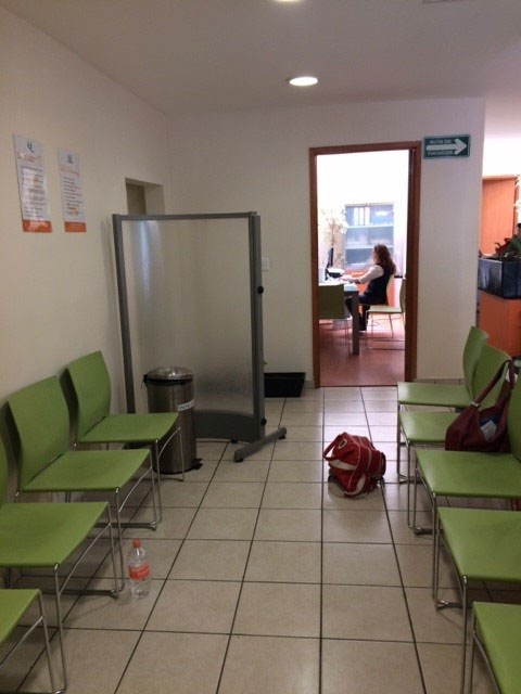 Medical facility waiting room with two rows of green chairs and a view into a medical worker's office