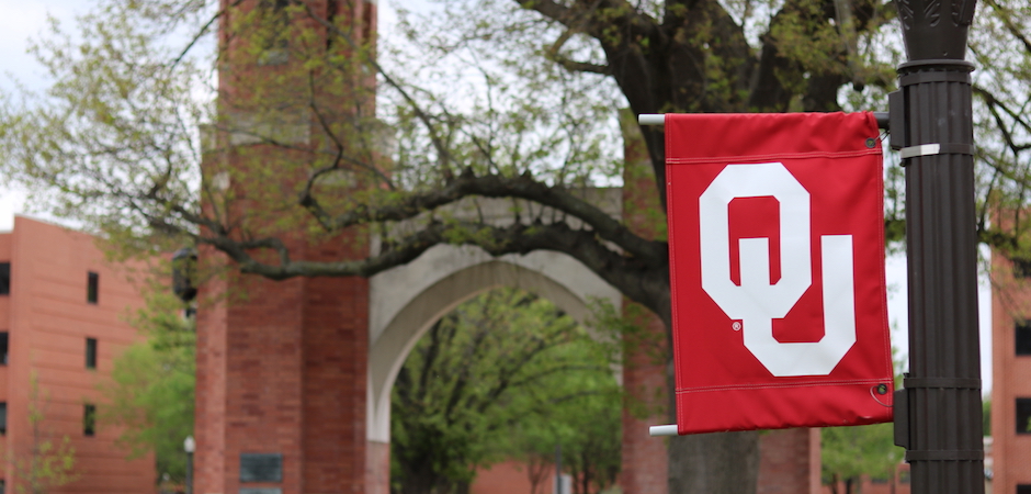  OU flag on a light pole with campus archway, Lindsey Street, and Cate Centers One and Two in background 