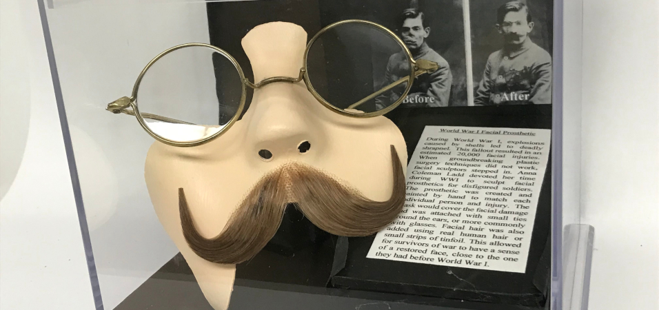 Photo of an exhibit of student work. The photo includes a mask that seems like a realistic, partial human face with glasses and a mustache. In the background are two photos, one labeled "Before" of a young man with part of his face disfigured; the other is the same young man wearing the mask. The explanatory text beneath the two photos is: "World War I Facial Prosthetic During World War I, explosions caused by shells led to deadly shrapnel. This fallout resulted in an estimated 20,000 facial injuries. When groundbreaking plastic surgery techniques did not work, facial sculptors stepped in. Anna Coleman Ladd devoted her time during WWI to sculpt facial prosthetics for disfigured soldiers. The prosthetic was created and painted by hand to match each individual person and injury. The mask would cover the facial damage and was attached with small ties around the ears, or more commonly with glasses. Facial hair was also added using real human hair or small strips of tinfoil. This allowed for survivors of war to have a sense of a restored face, close to the one they had before World War I."
