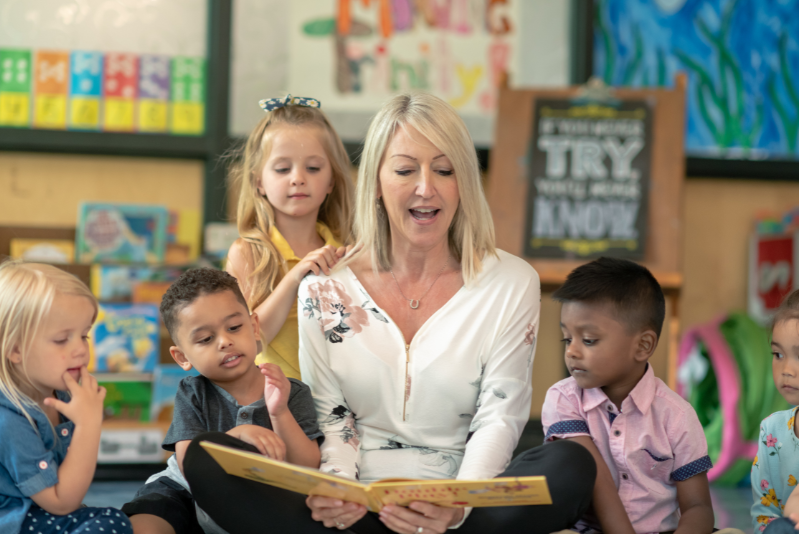 blonde woman in a white shirt sitting on the floor with a book in hand reading to students who are sitting around her