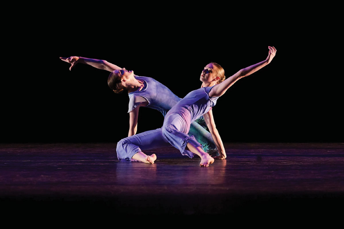 Two dancers bending their backs and extending their arms and legs, while sitting on a darkened stage.