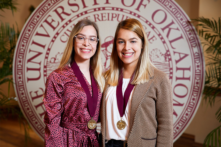 Elizabeth and Sarah Cermak pose wearing bronze medallions in front of a large OU Seal backdrop