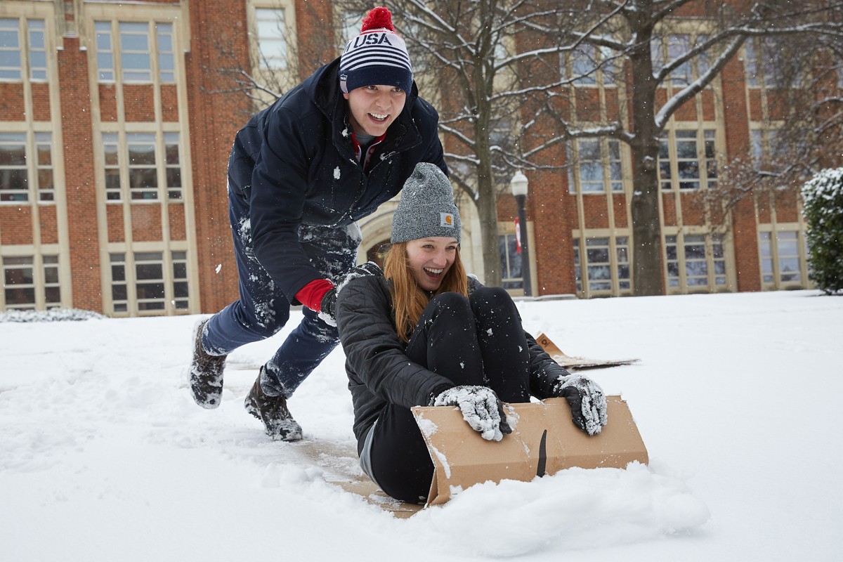 One student pushes another through the snow on a makeshift, cadboard sled