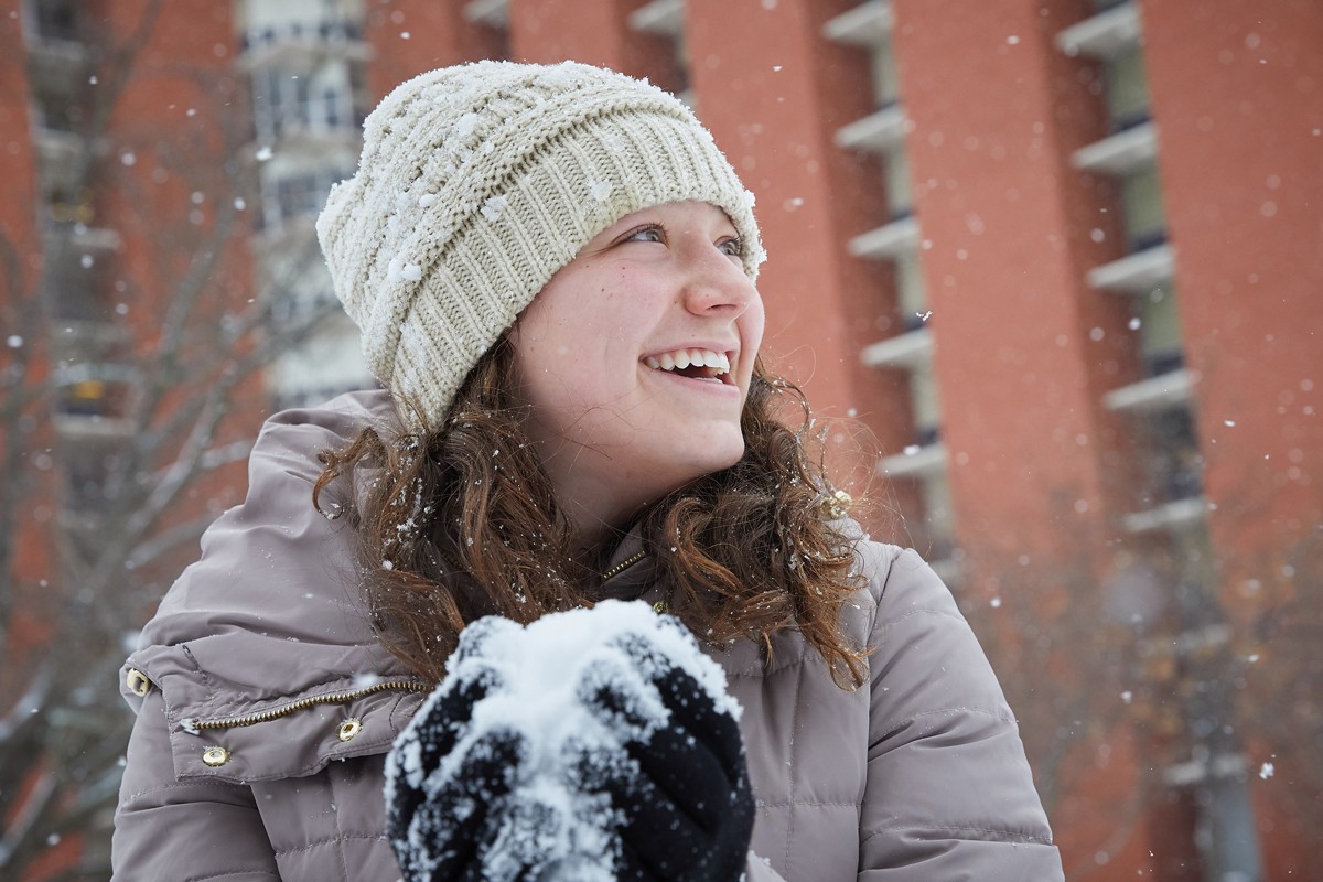 Student packs a snowball in gloved hands outside near dorms