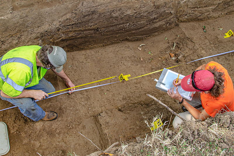 Two people photographed from above, in a dirt channel, using measurig equipment