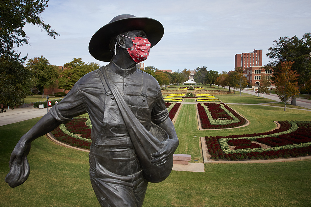 The Sower Statue is wearing a fabric PPE mask with an OU pattern, the planted flowerbeds stretch out behind it.