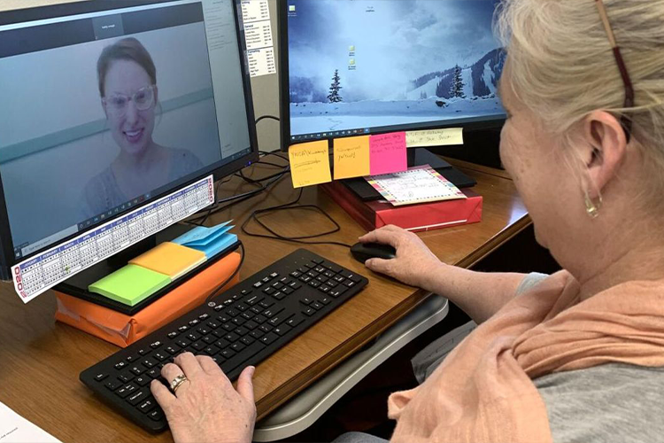 Woman sits at a desk, at a computer, engaged in a video call with a second woman.