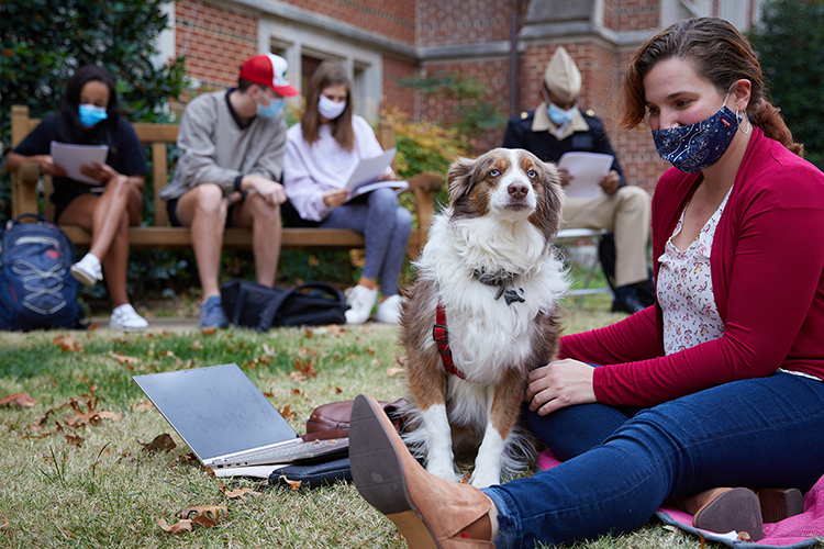 Student studies on the North Oval lawn, a dog with her. More students can be seen, out of focus in the background.