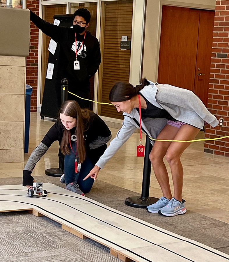 Three students pace a small robotic vehicle on a wooden track