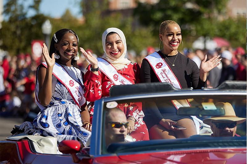Three female students sit in the back of a car waving during the homecoming parade