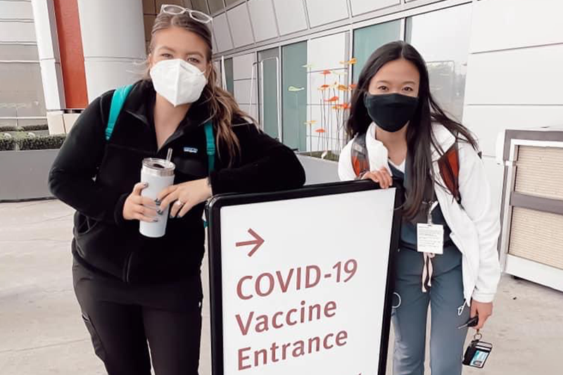 Two masked students stand near a "COVID Vaccine Entrance" sign