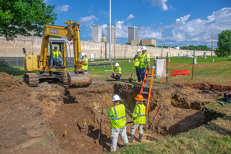 Workers in hard hats and high visibility vests work excavating at Oaklawn cemetery