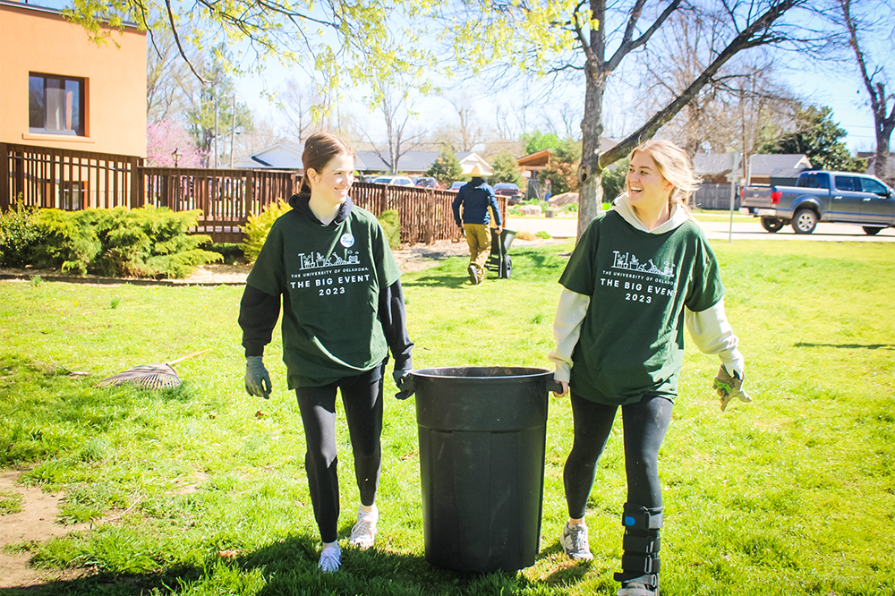 Two female students carry a trashcan together across a yard.