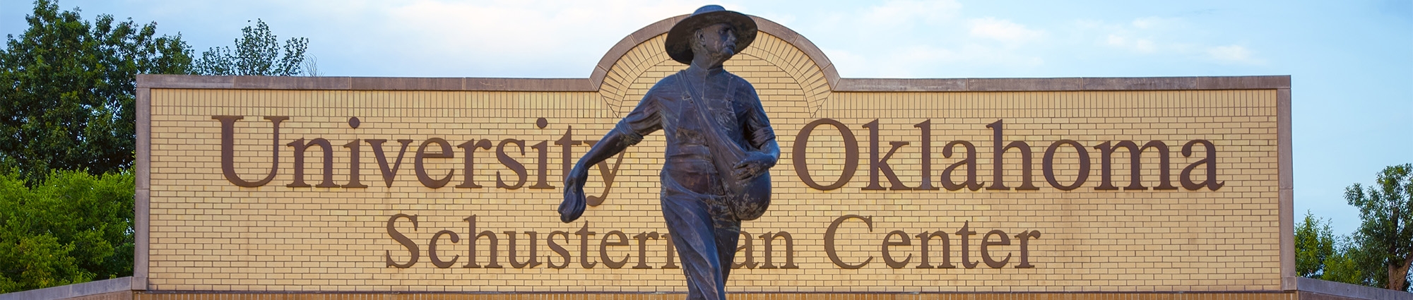 A statue of the OU Seed Sower, a figure in a wide brimmed hat with an arm extended behind him. In the background, a brick sign for the University of Oklahoma Schusterman Center.