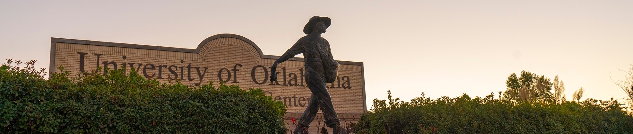 The OU-Tulsa seed sower statue, standing in front a sign featuring the name of the university, with the sun setting in the background