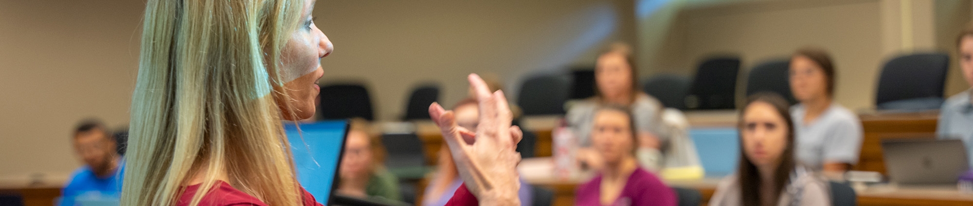 A professor speaks to a class, gesturing with both hands as students in the background look on
