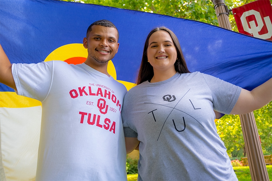 Two smiling students in OU-Tulsa tshirt hold a Tulsa flag aloft above their head. An OU light post is visible in the background.