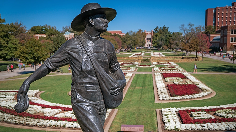 A photo of the Seed Sower statue at OU in Norman, with the green South Oval stretching behind it, festooned with red and white flowers 