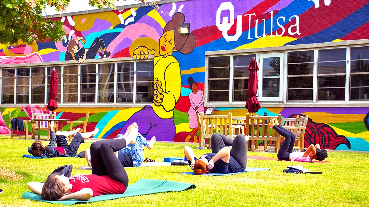 OU-Tulsa students perform a yoga pose in the grass in front of a large, colorful mural on the OU-Tulsa campus