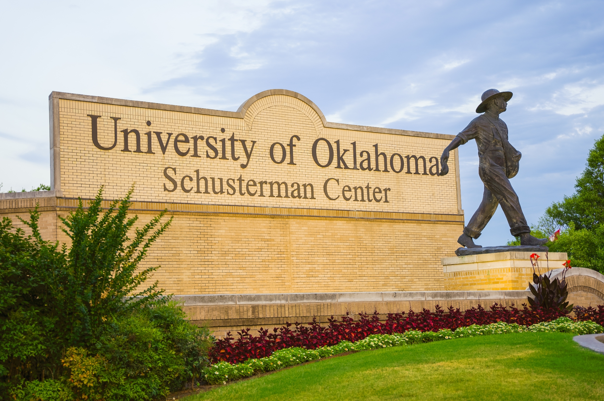 A sign on blonde brick reading "OU-Tulsa Schusterman Center" with a statue of the OU Seed Sower in front. A blue sky stretches above.