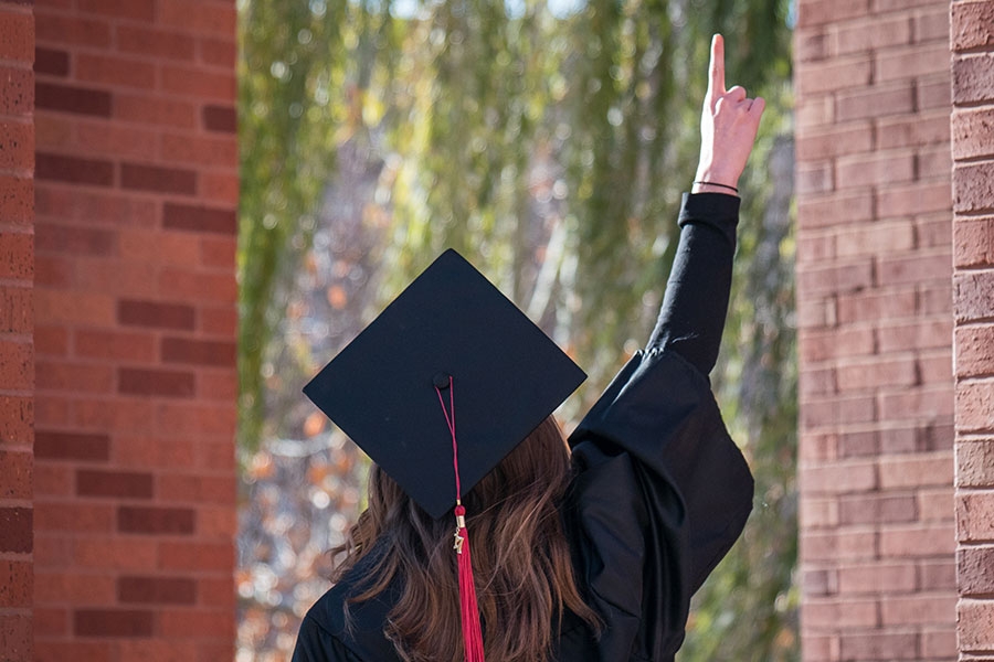 Student wearing cap and gown holding up symbolic index finger under Bizzell Clocktower. 