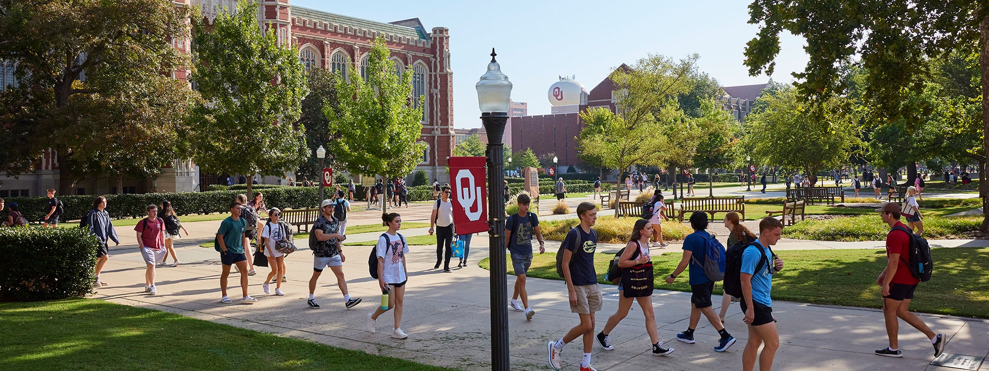 Students walking on the University of Oklahoma campus, with an OU flag and lamppost in the foreground.