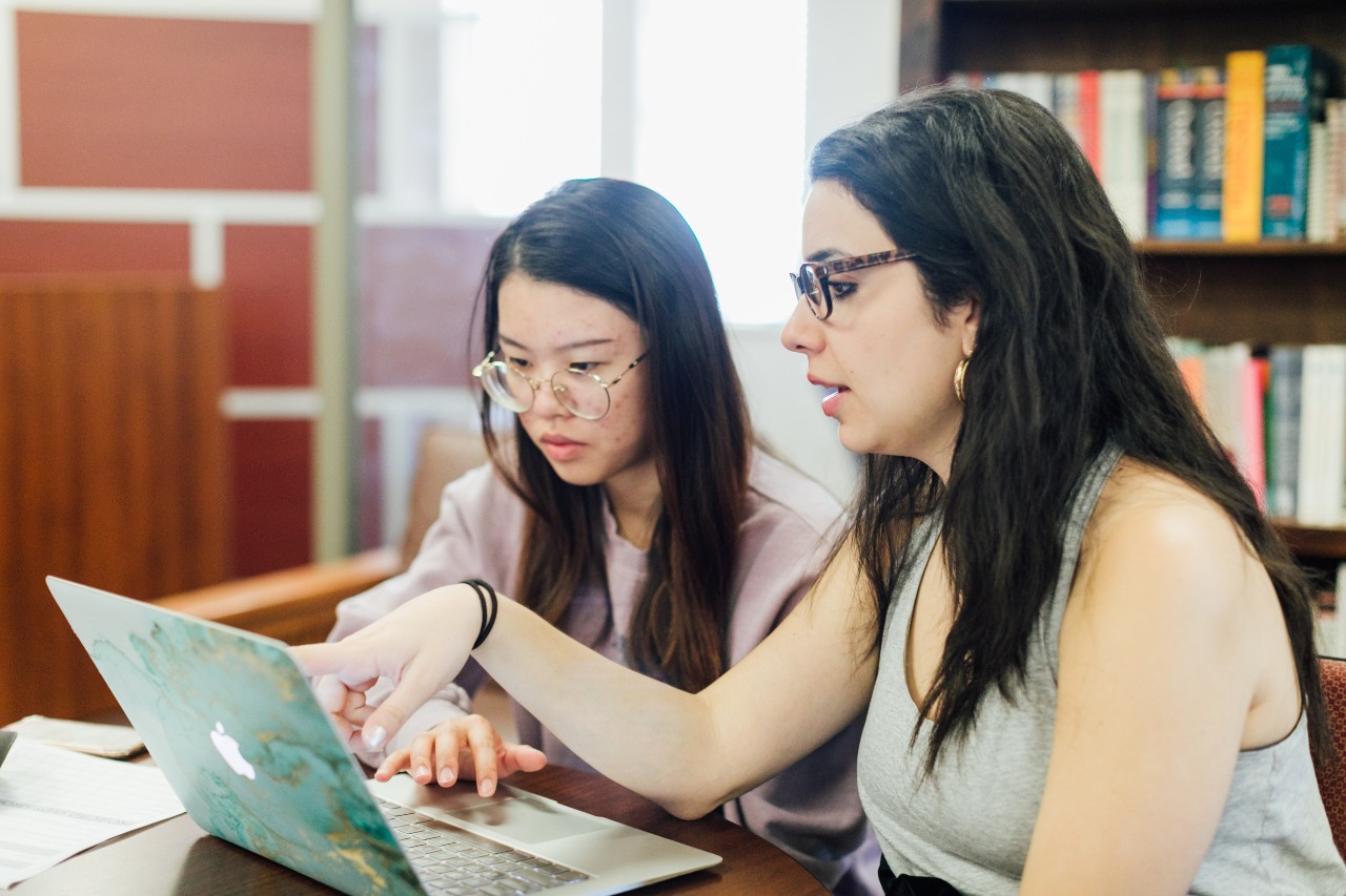 Two students with long dark hair sit next to each other at a table in a tutoring center, both focused on the open laptop in front of them. One student gestures with her hand, while the other listens attentively. Both are seated on chairs, surrounded by a quiet academic environment, with papers and study materials spread out on the table.