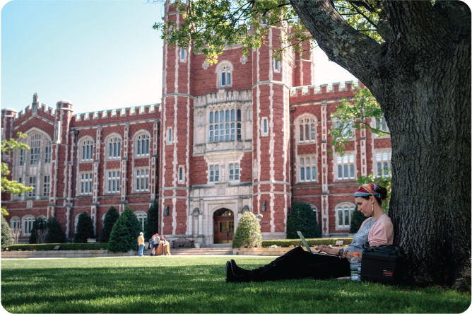 A student leaning on a tree and working on her laptop on the lawn in front of the library. 