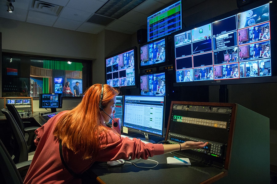 A woman in a control room with broadcasting equipment looking at multiple screens. 