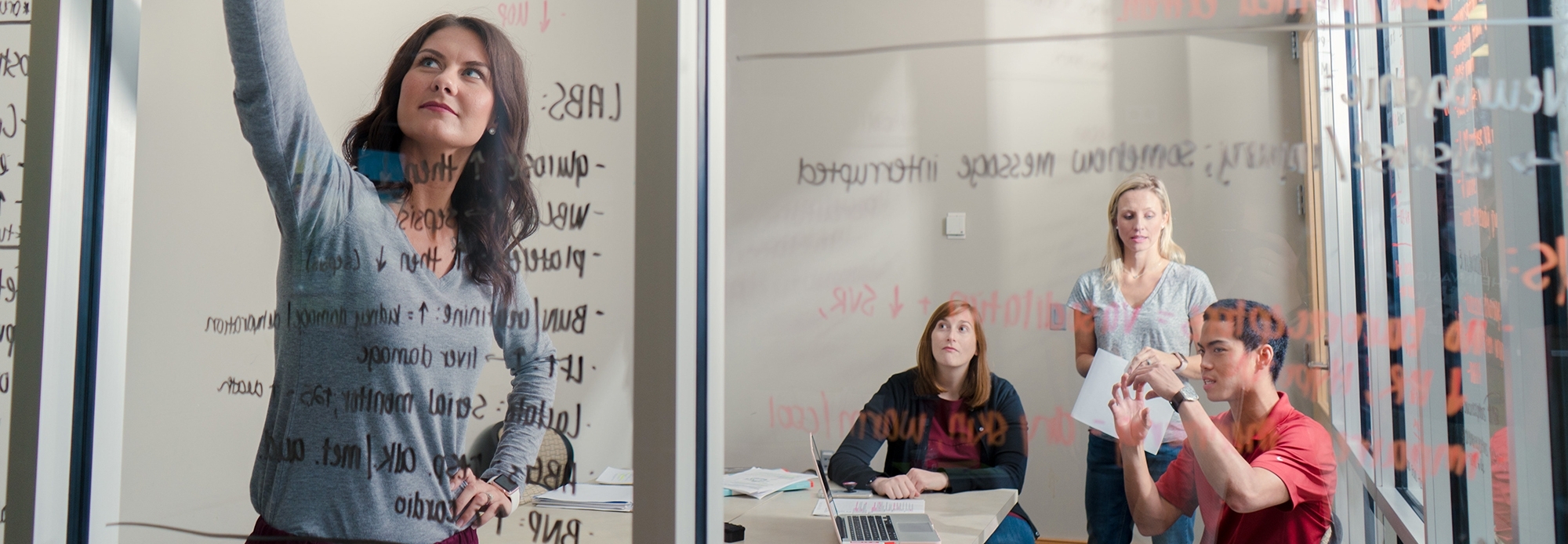 An person writing on a whiteboard, with three people in the background.