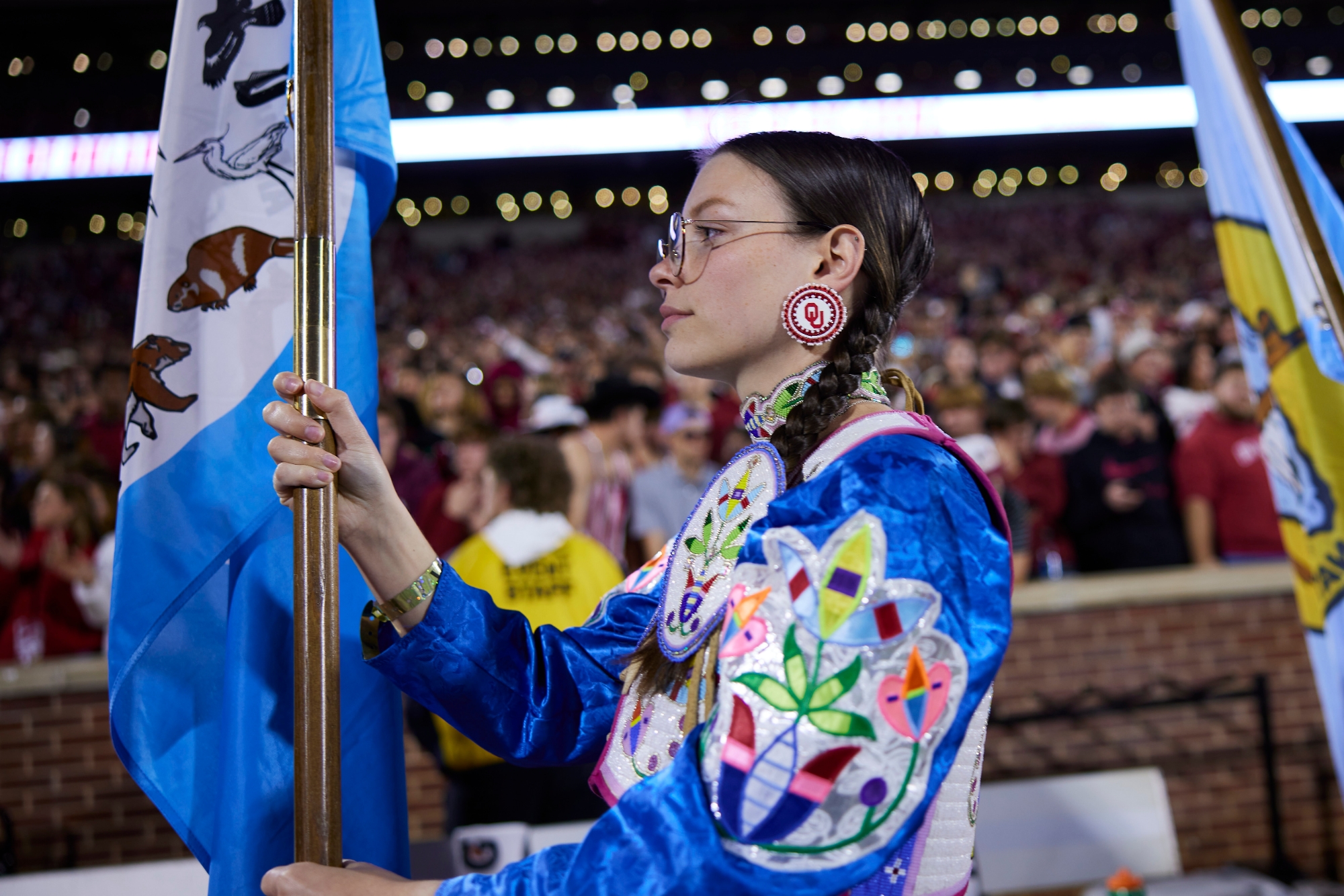 An OU student in full regalia holding a flag as she walks through the field