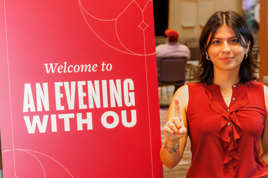 Current OU student holding up a one next to a sign that says "Welcome to An Evening With OU"
