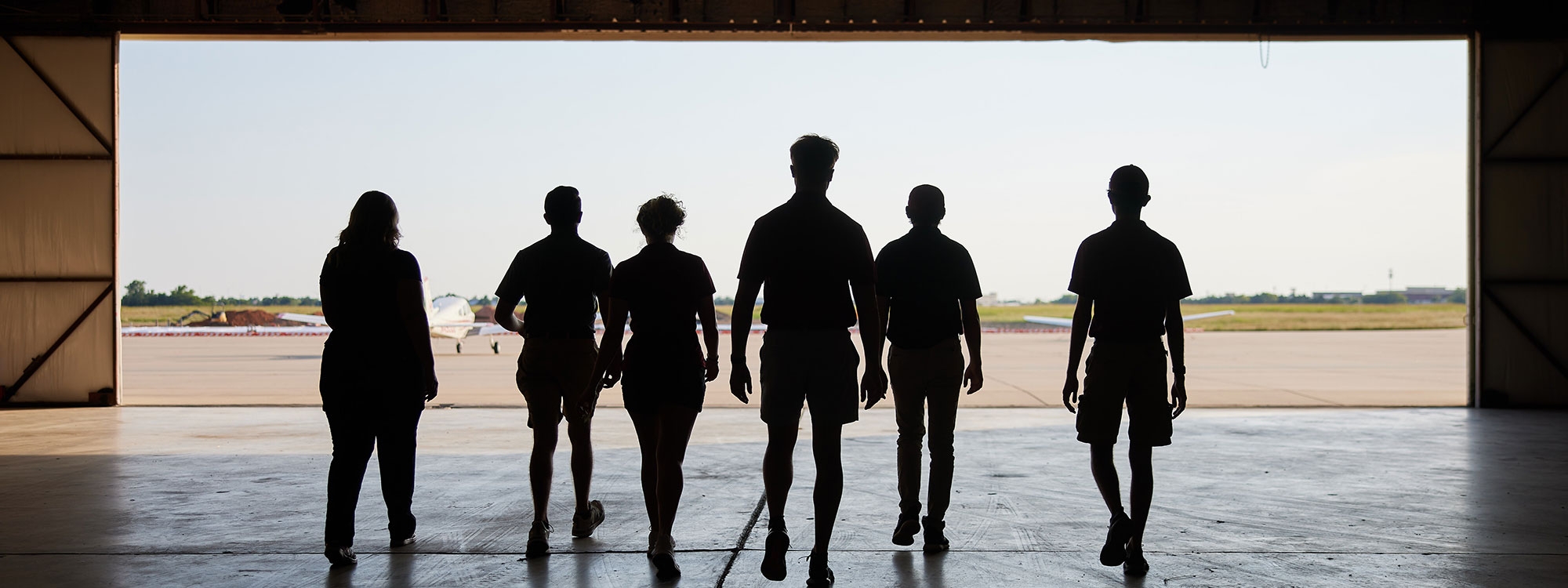 A group of backlit students walking out of an aircraft hangar.