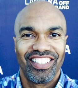 A headshot of Dr. Buckmire, smiling, in front of a blue background. L, Off, And, A.