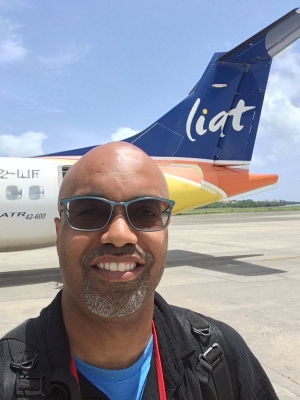 Dr. Ron Buckmire stands in front of an airplane on a runway, surrounded by blue sky and cumulus clouds. Liat.