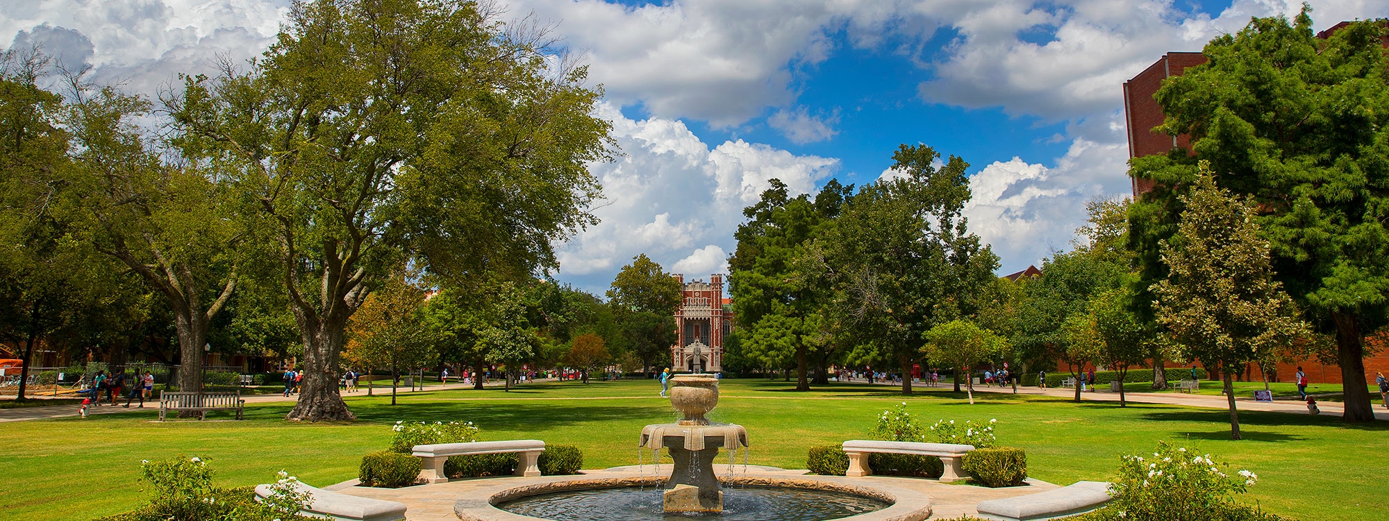 Trees and buildings on OU's campus, with a blue sky and white clouds in the background and a fountain and benches in the foreground.