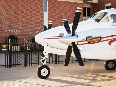 A white airplane parked outside the Airport terminal with the University of Oklahoma Displayed on the building.