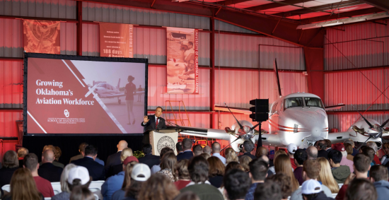 Aviation Hangar with a King air airplane in the cornor. President HArroz is at the podium addressing the Aviation program to a hangar full of attendees.