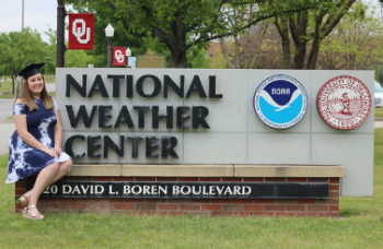Elizabeth Leslie sits on the edge of the National Weather Center sign, wearing her graduation cap. OU, National Weather Center, NOAA, 120 David L. Boren Boulevard.