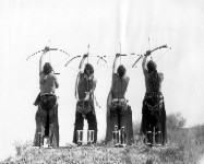 Four Indigenous men shooting bows up into the sky, in black and white.