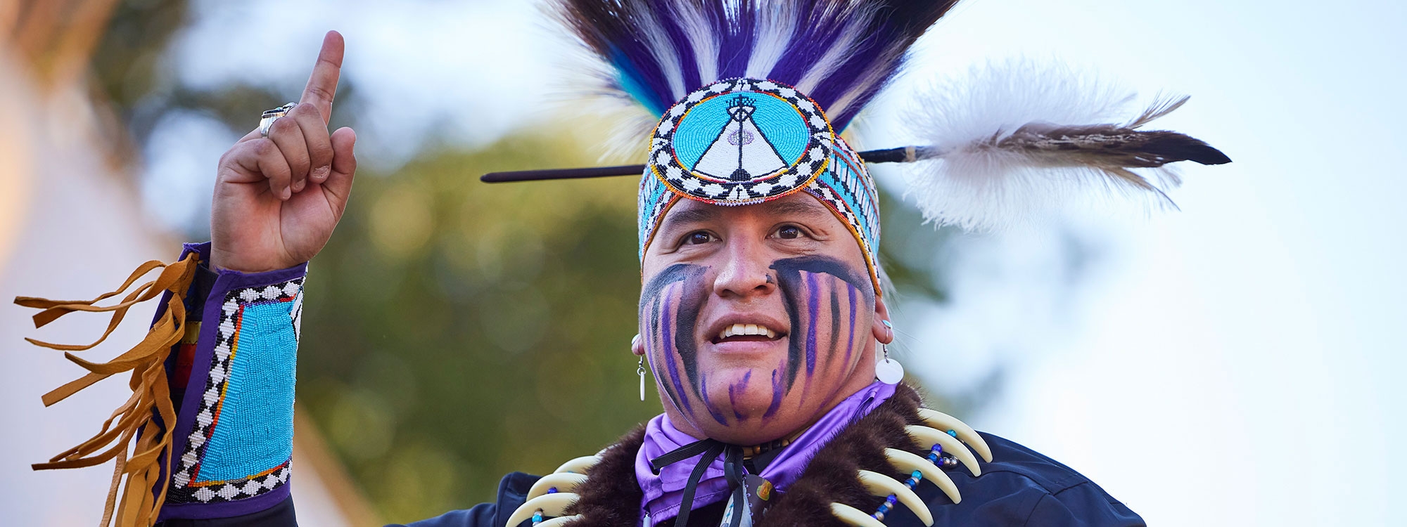 A man in purple, blue, and white Native clothing, raising his pointer finger.