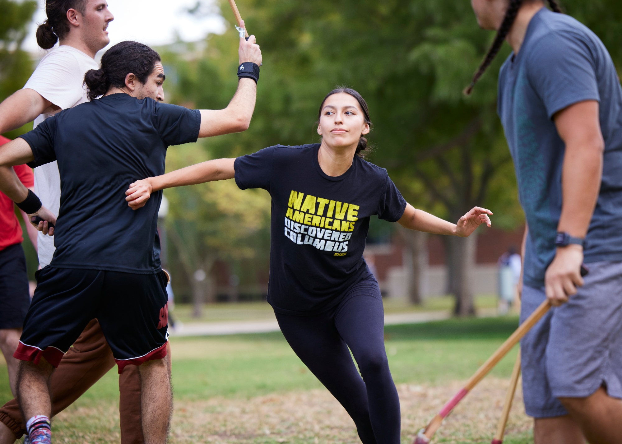 Four Native students playing stickball. A young woman is in the middle pushing her way through with a shirt that reads "Native Americans Discovered Columbus."