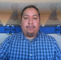 A picture of Dane Poolaw. He is in a chile pepper field and holding a basket of chile peppers in a mask.