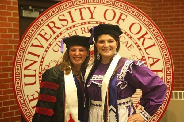 Picture of two women standing in front of the University of Oklahoma seal. They are both wearing graduation caps and the women on the right is wearing a purple traditional dress, (possibly Cherokee.)