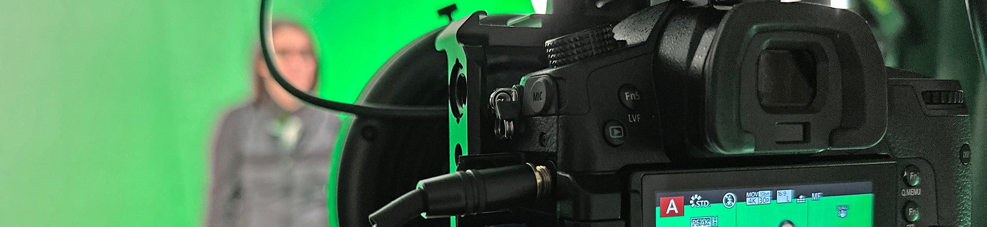 Photo of video studio camera in the foreground. The background is blurry with a person standing in front of a green screen.
