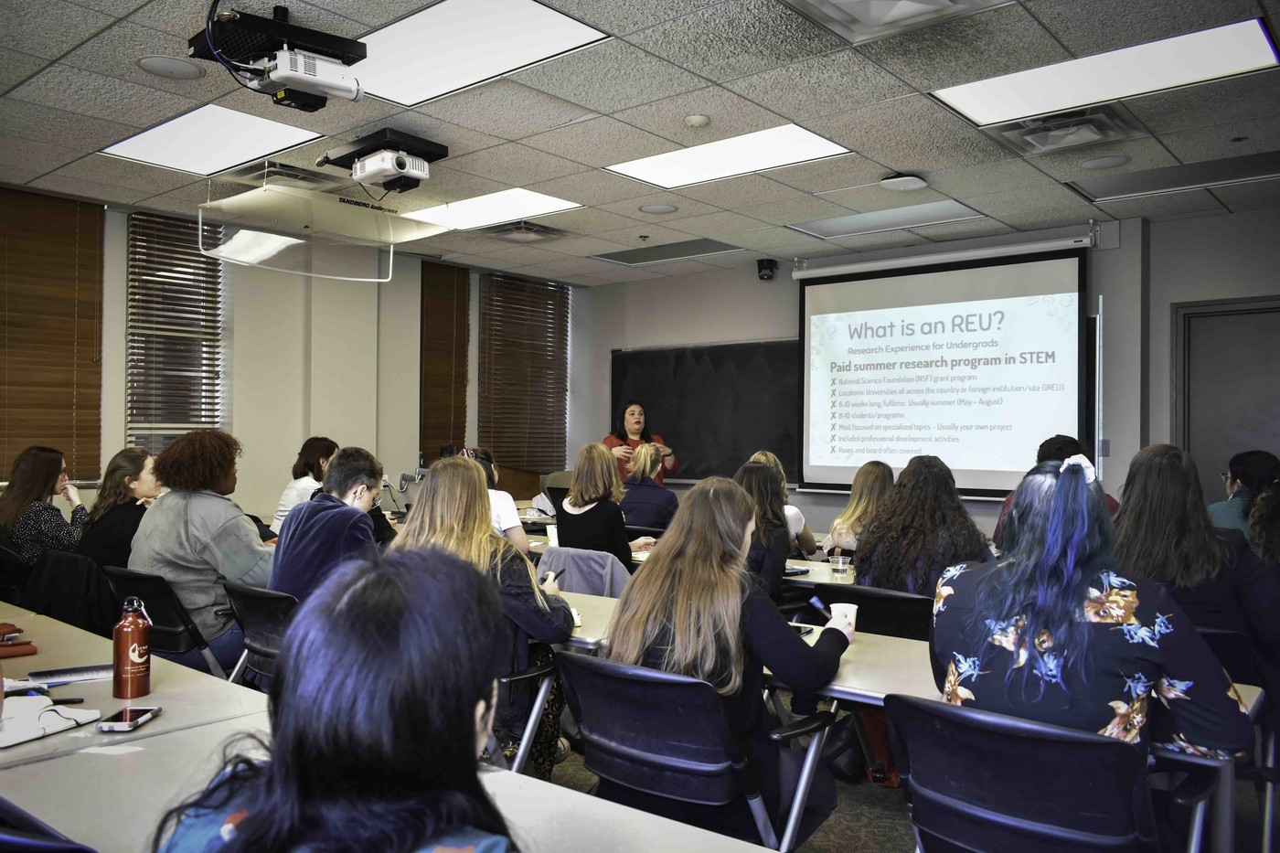 People sit at desks in on of the physics classrooms watching a presentation on the projector screen.