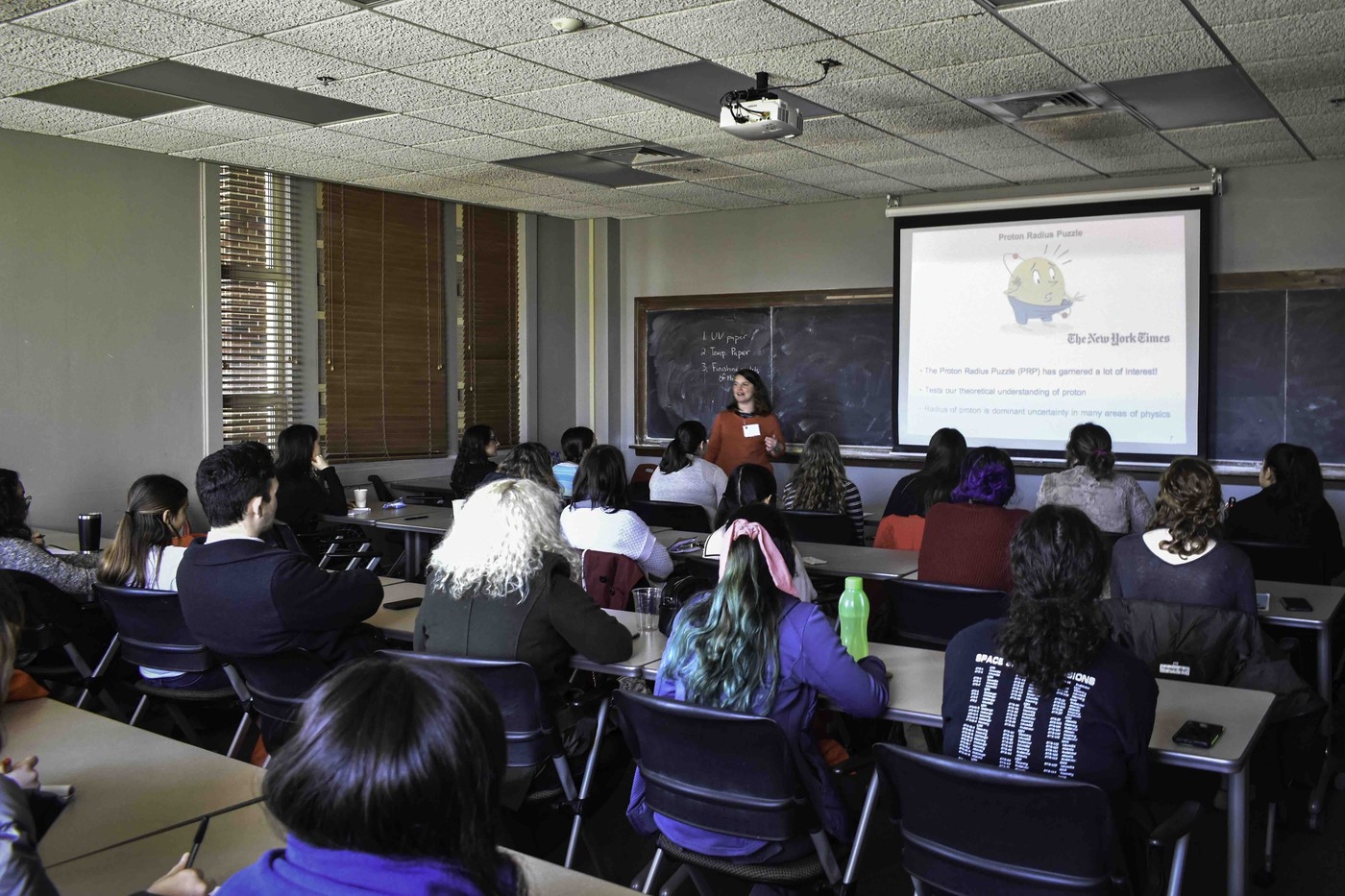 People sit at desks in on of the physics classrooms watching a presentation on the projector screen.