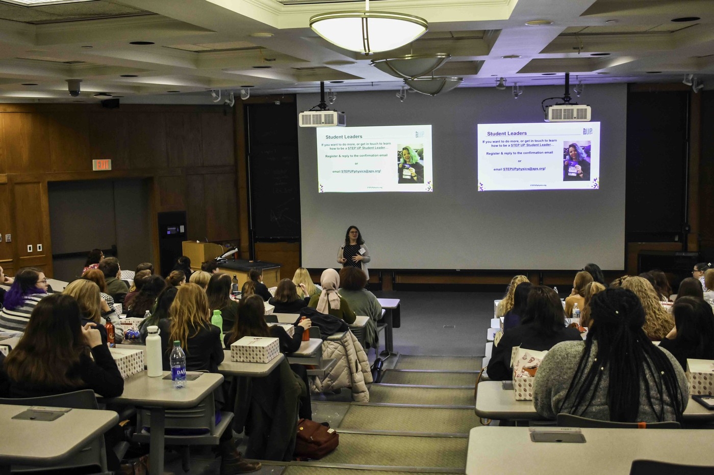 A group of attendees sits in a large lecture hall to watch a presentation.